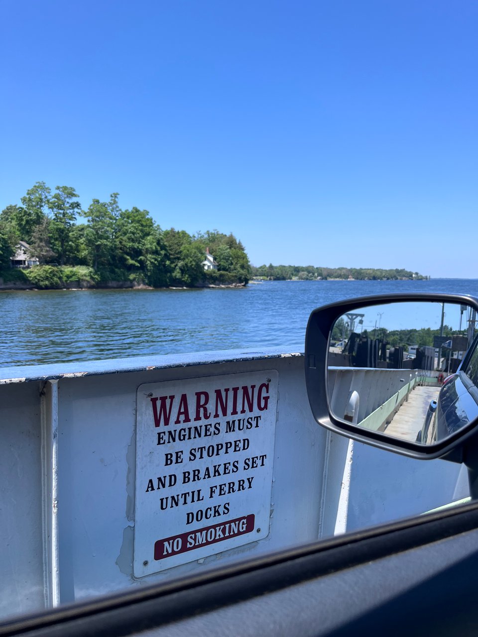 A color photograph of the perspective of a driver on a ferry looking out the window of their car. The sign on the side of the ferry says "WARNING: ENGINES MUST BE STOPPED AND BRAKES SET UNTIL FERRY DOCKS. NO SMOKING"