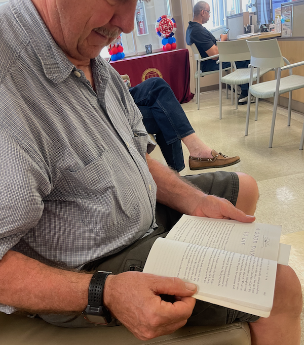 A white man with a mustache wearing a watch on his right wrist holds a book open to a chapter titled "A Good Day to Die"