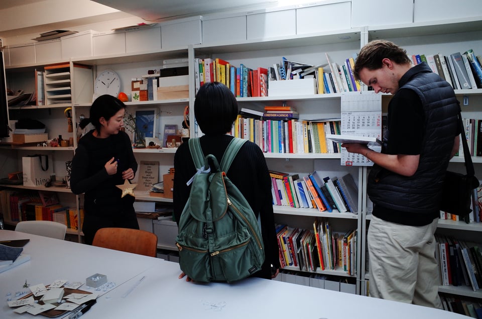 Three people stand in front of floor to ceiling bookshelves, and behind a table with several star drawings on top of it.
