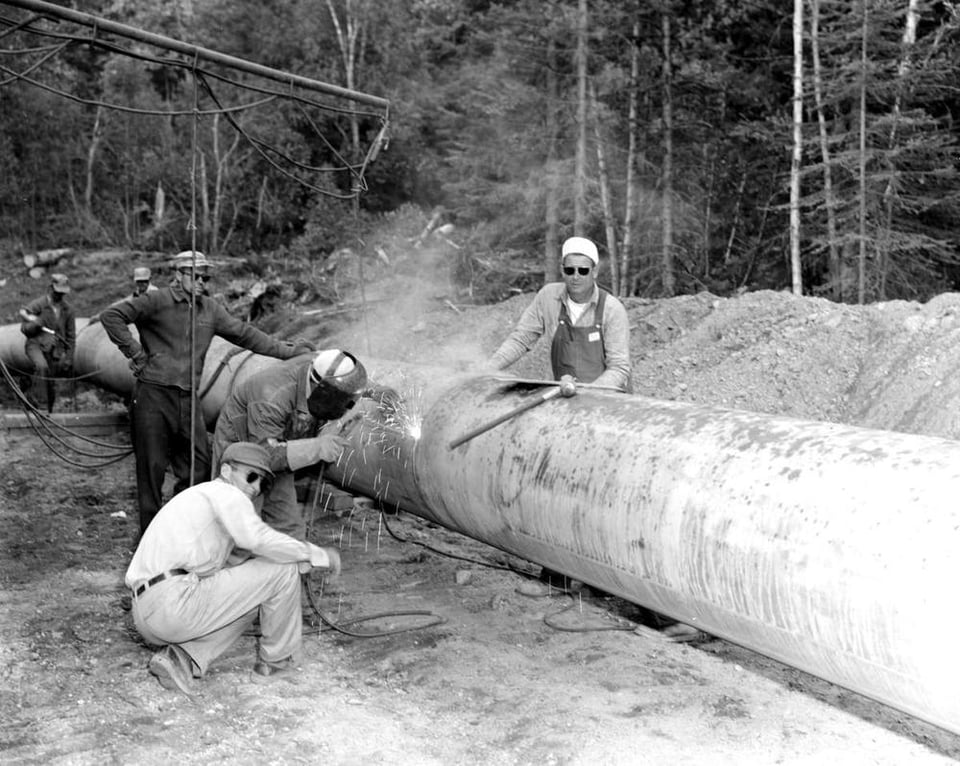 black and white photo of men standing around a pipeline. One is welding and the others are standing around.