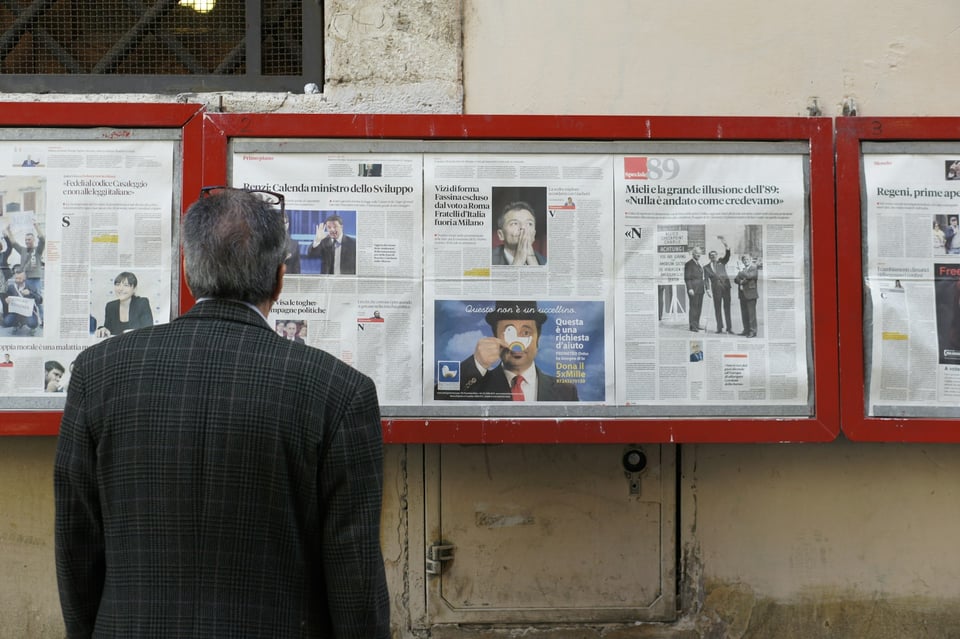 A person stands in front of newspapers on display, in cases on a wall. Photo by Filip Mishevski on Unsplash.