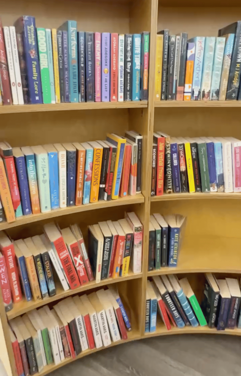 Curved wooden bookshelves filled with rows of colourful books, including a mix of contemporary fiction and other titles.