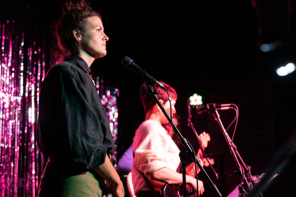 Photograph from the live performance that Alive People by Ruth Garbus was recorded from. The image shows Julia Tadlock in the foreground at a microphone and Ruth Garbus playing guitar next to her, and Julie Bodian in the background.