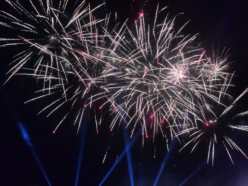 Feuerwerk. Weiße Leuchtspuren vor Nachthimmel, dazu nach oben strebende blaue Scheinwerferstrahlen.