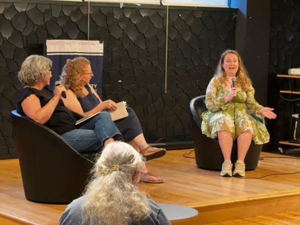 Two women sit on black seats and hold books and one has a microphone. Across from them, a young woman in a green and white dress speaks into her microphone and is gesturing.