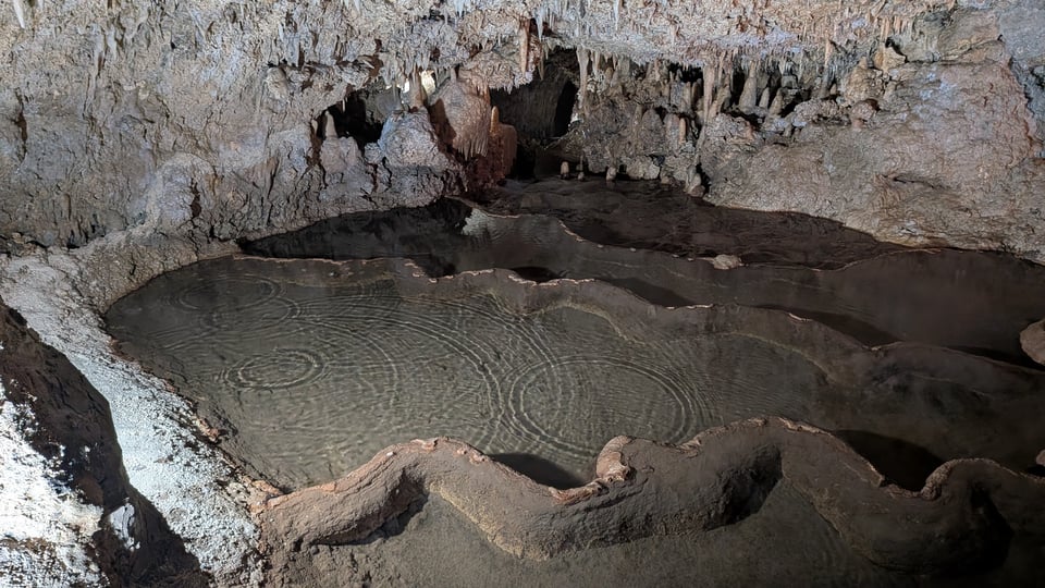 A series of shallow pools in a grey and brown cave, illuminated by soft white light. Water is falling from the ceiling and casting the shadow of ripples into the pools.