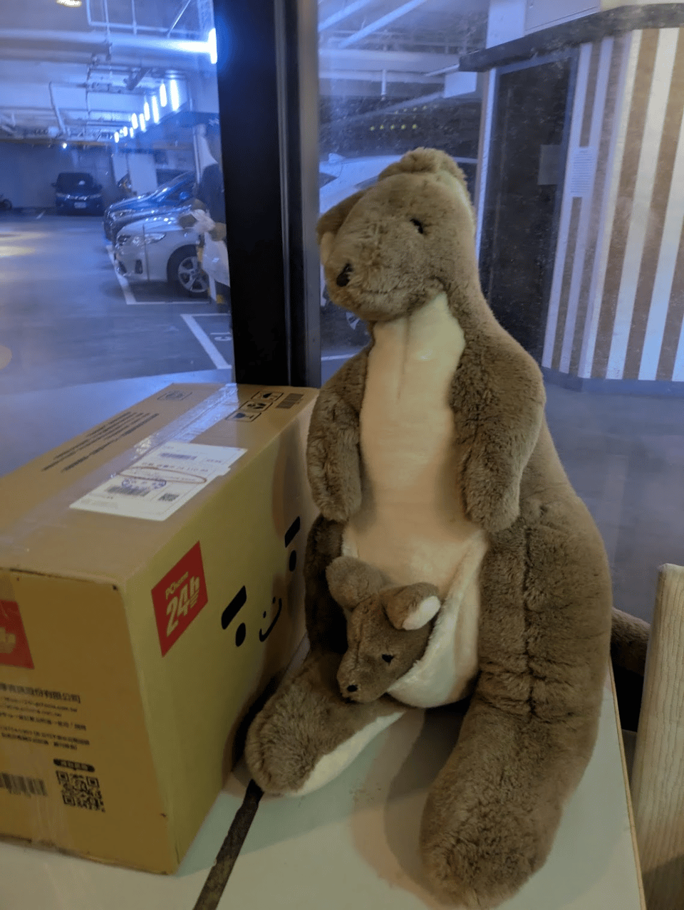 A large stuffed kangaroo with a little roo in the pouch. It's sitting on top of a table next to a large package, with an underground garage in the background.