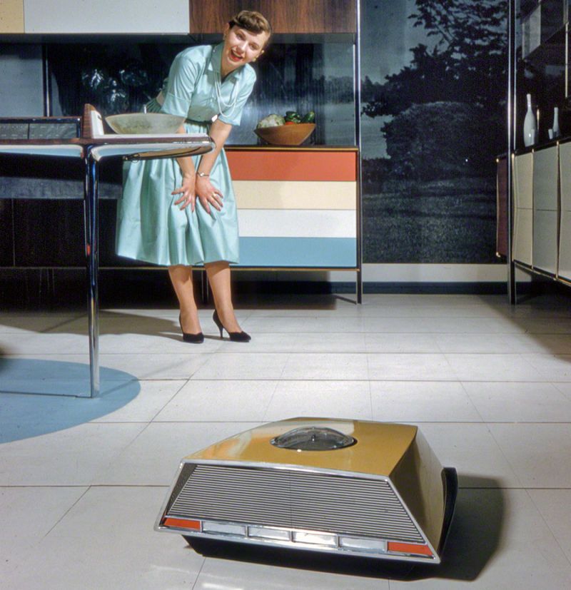 An ad for the Miracle Kitchen of the Future from RCA/Whirlpool. A stereotypical 50s housewife in a blue dress and pearls with black heels looks across a dining area at a device that looks like a cross between an Oldsmobile and a Roomba in a mustard yellow chassis. She's utterly charmed. It's wondering what there is to vacuum in this pristine room.