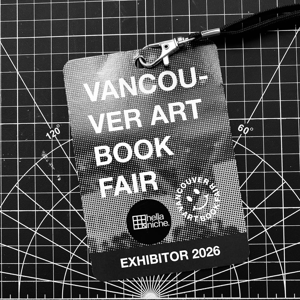 A black and white photograph of a VABF Exhibitor 2026 lanyard badge resting on a cutting mat. The badge displays "Vancouver Art Book Fair" in large white type with a halftone gradient, and bears a hellaniche sticker in the lower centre.