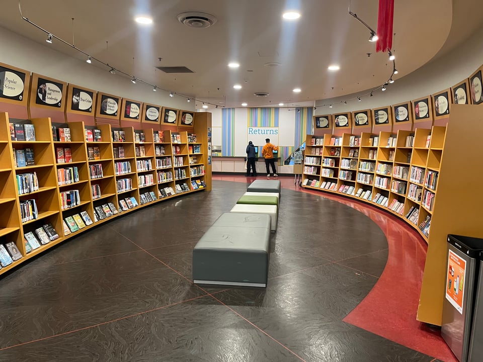 An area of the library with gently curving shelves on the side, and some soft cushioned-benches in the middle. A library desk with a “Returns” sign is visible beyond the end of the shelves.