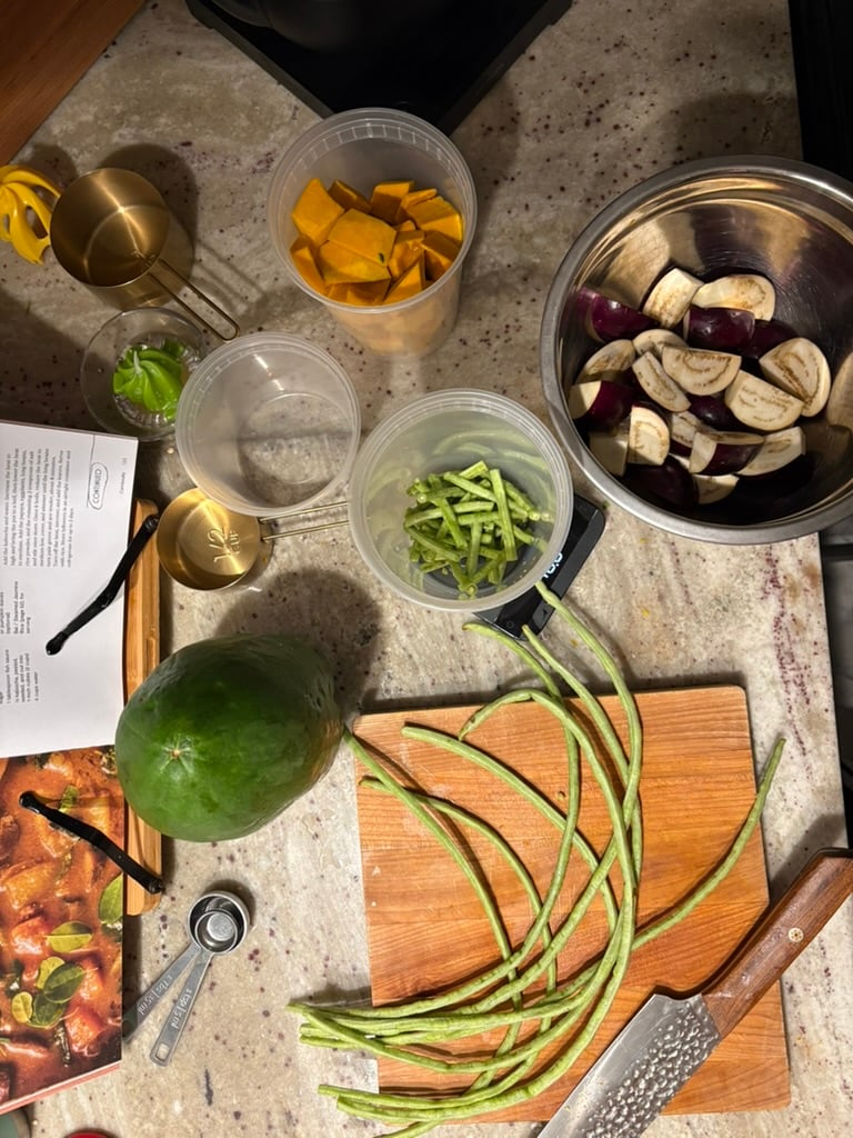 Green papaya, long beans, baby eggplant, and kabocha squash laid out for preparation on a cutting board with a nakiri.