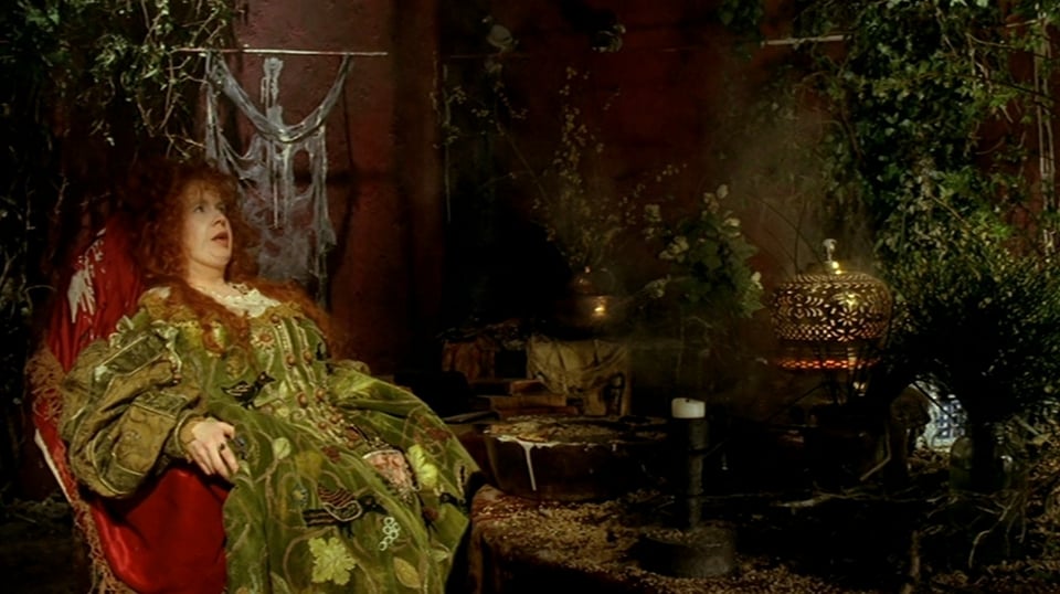 A woman with curly red hair wearing a light green dress covered in embroidery slouches in a chair draped in red. The room she is in is covered with trailing vines and cobwebs.