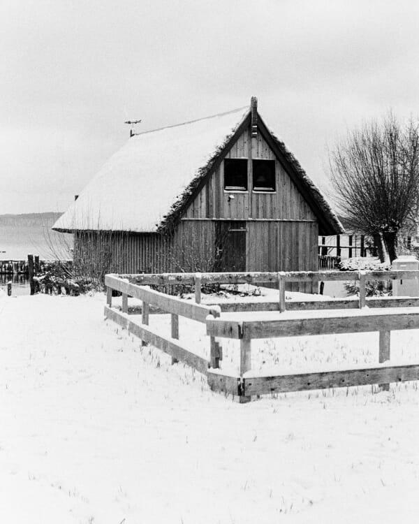 B&W film photo of a thatched-roof hut behind a wooden fence, surrounded by a pristine field of snow.