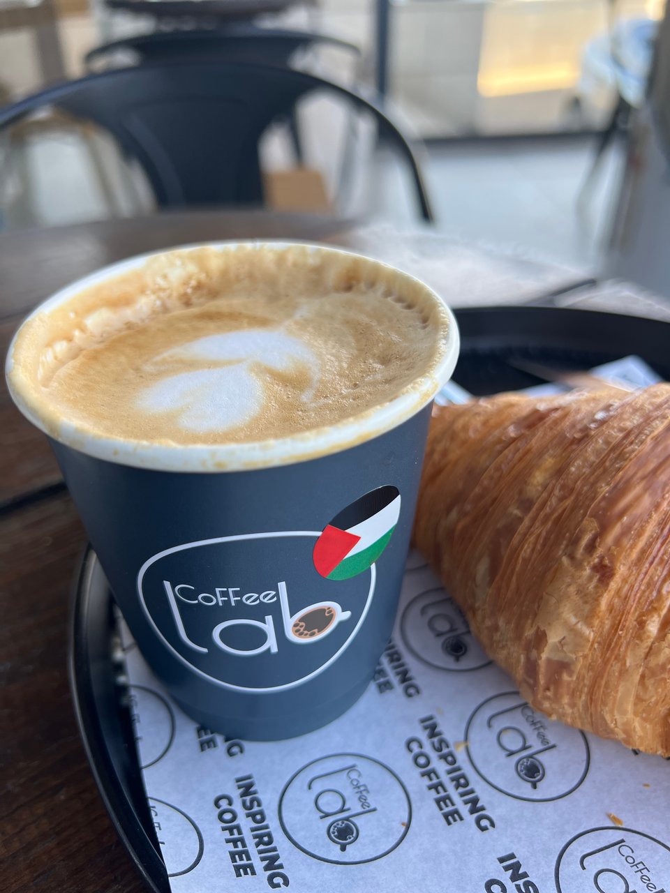 A coffee cup with the logo of Coffee Lab and a decal of a Palestinian flag, on a plate with a croissant