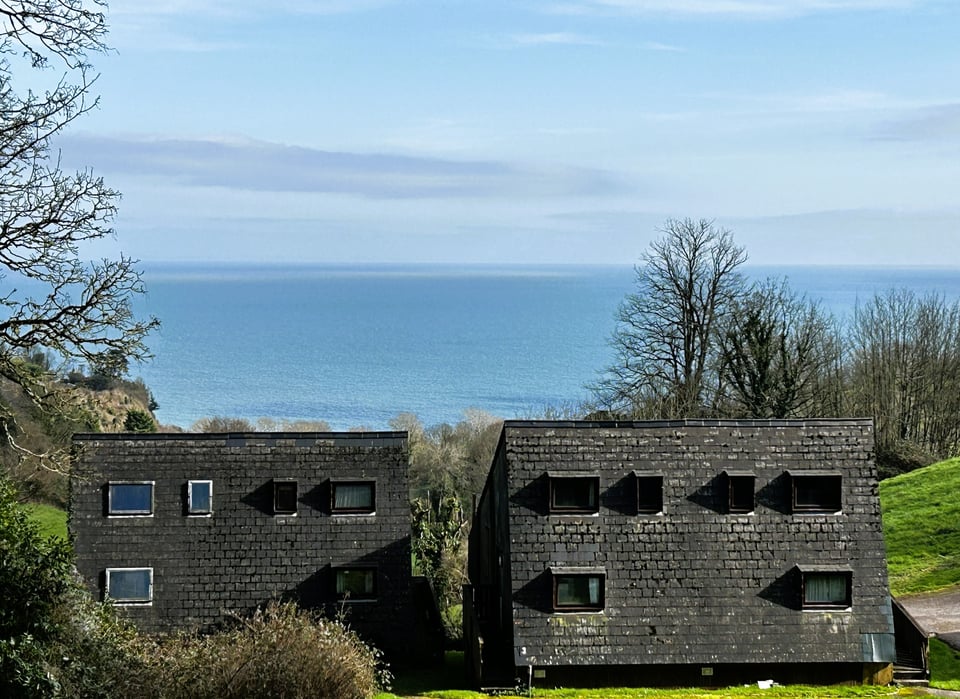 Two pairs of lodges from the rear, looking out towards a wide blue sea. The lodge's roofs drop down almost to the ground. The one on the furthest left has original flat skylights. The remainder have hooded dormer windows that have been set into the slate.