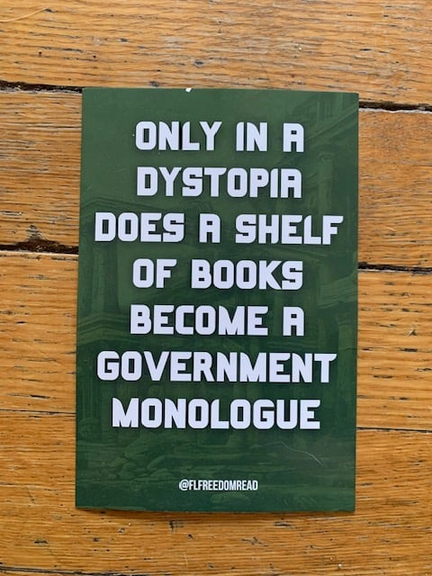 Image of a green postcard on a wood floor. The postcard has white text that reads "Only in a dystopia does a shelf of books become a government monologue," with the handle for the Florida Freedom to Read Project (@Flfreedomread) below it.