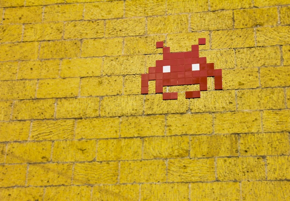 A photo of a yellow brick wall, and in the upper right quarter there is an image of an alien from the original space invaders game, depicted in red and white tiles.