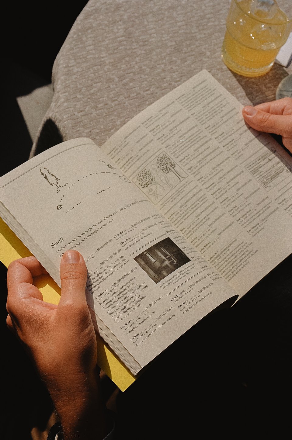 [A pair of hands holds the phone book open to a six column spread of entries peppered with illustrations and images. Sunlight hits the small round table behind it, where a yellow drink sits.]