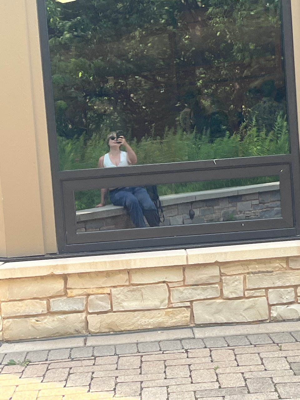 a reflection of Nola in a window with trees and grasses in the background. she is wearing a white vest, blue pants, and black sunglasses.