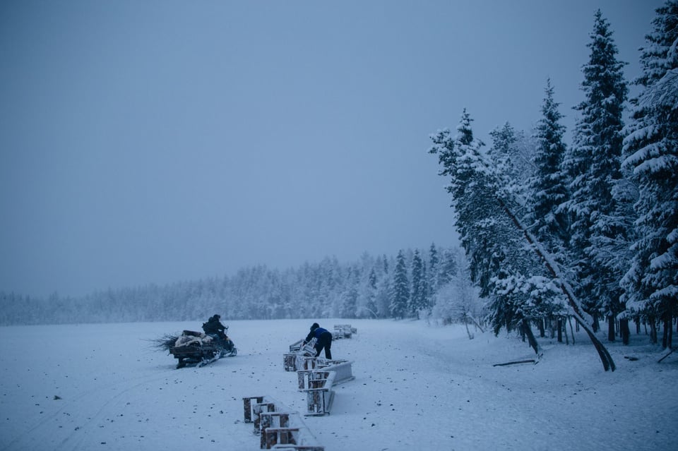 Thick snow blankets the landscape, trees coated in white, as a man sets up containers in the snow.