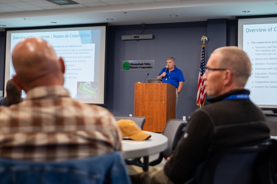 Tony Banks from VAFAIRS stands with a microphone in a blue shirt with a Powerpoint projection on cooperative development