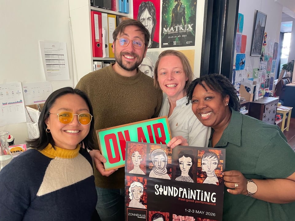 4 smiley people posing together, with a poster and an 'On Air' sign