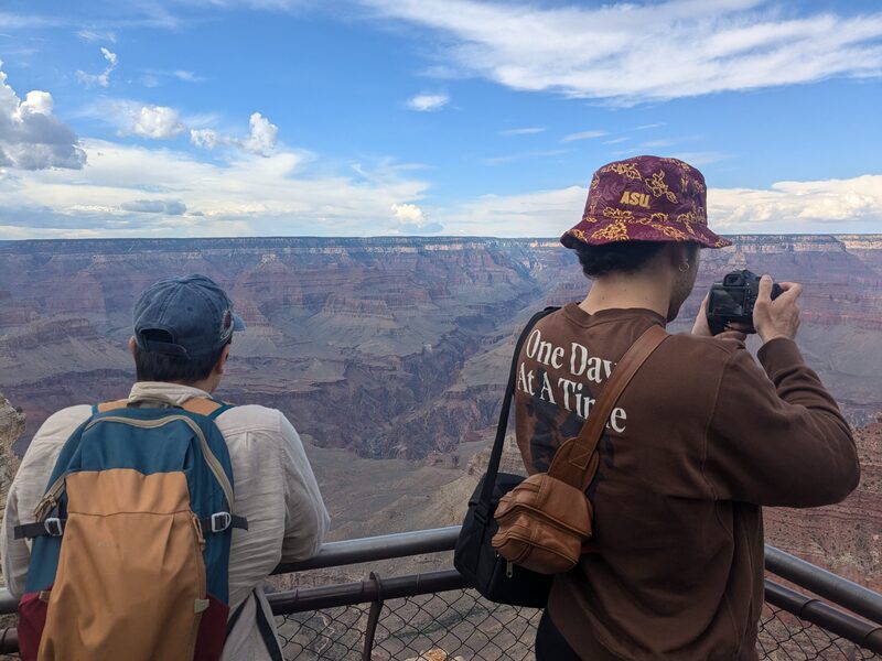 Two men facing the Grand Canyon. The taller of the two is on the right, taking a photo with a DSLR