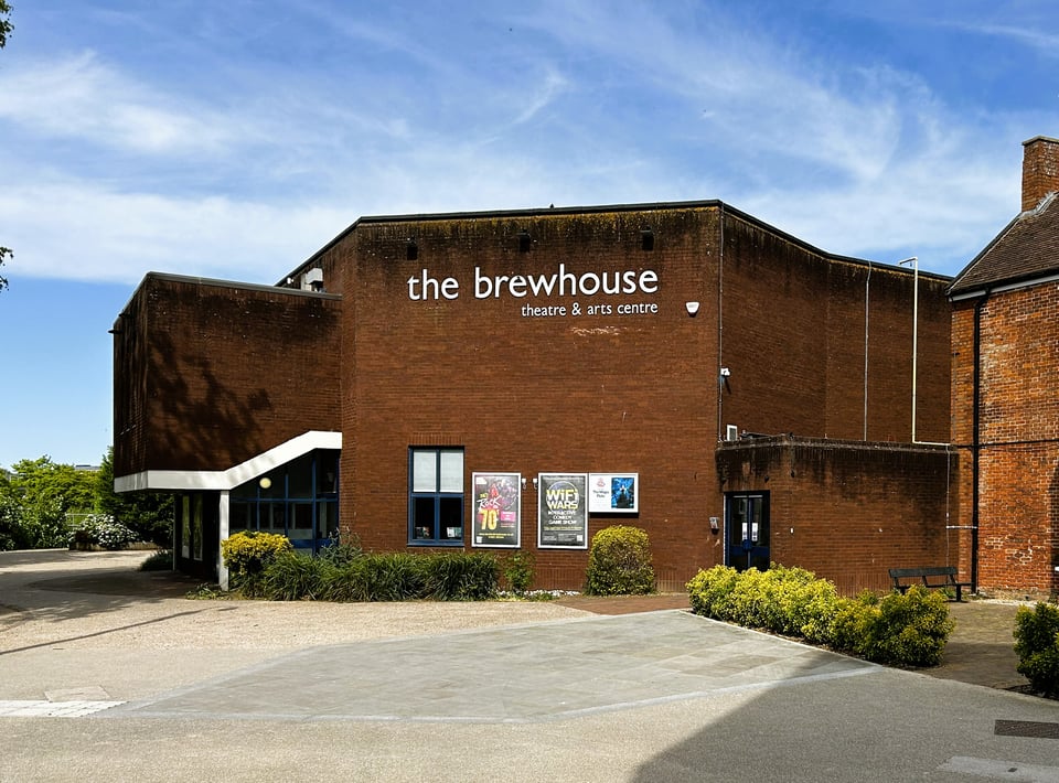 An octagonal brick theatre building. To one side, a room extends outwards on a concrete support. All of it is rather dark, and the brickwork needs a powerwash.