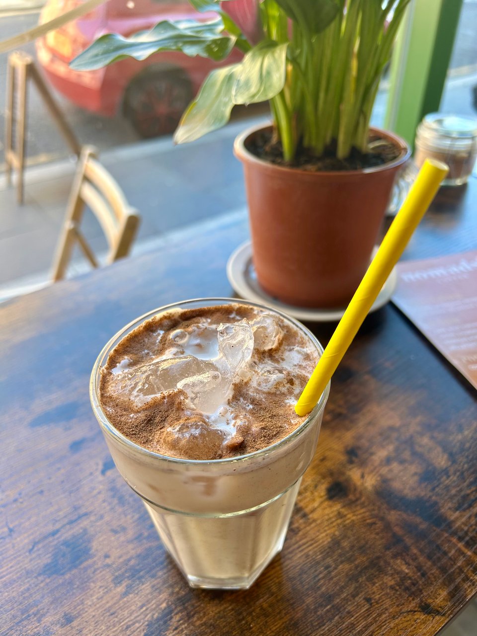 A chunky glass tumbler with a bright yellow straw is filled with an iced chai latte. It's standing on a polished and slightly scorched wooden table by a window. There's a bright green potted plant behind it. Image by Rowan Ambrose.
