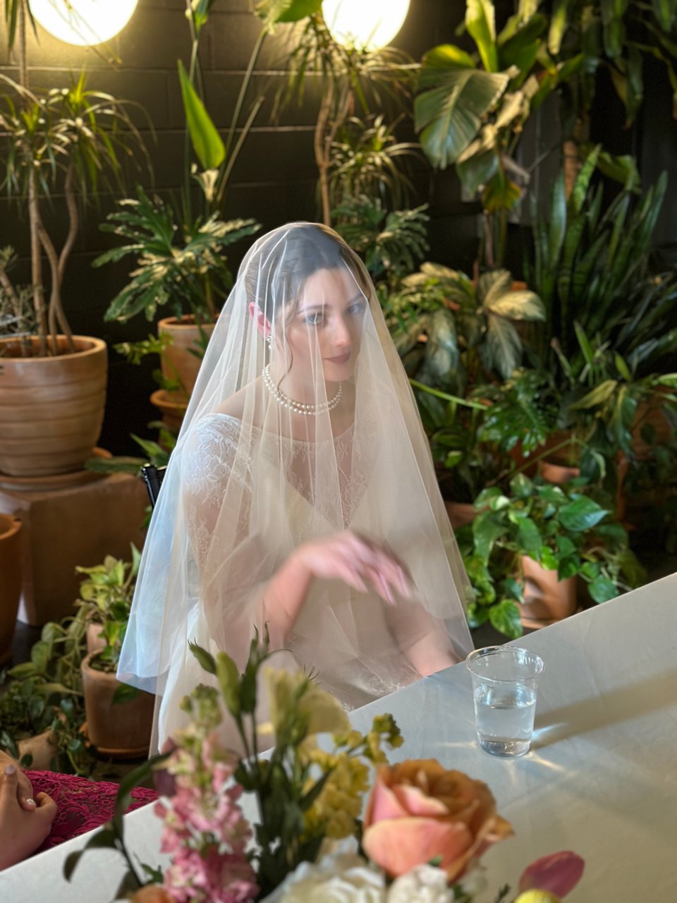 photo of white woman in bridal gown and veil, sitting at a table with flowers in the foreground and plants behind her. She's very beautiful.