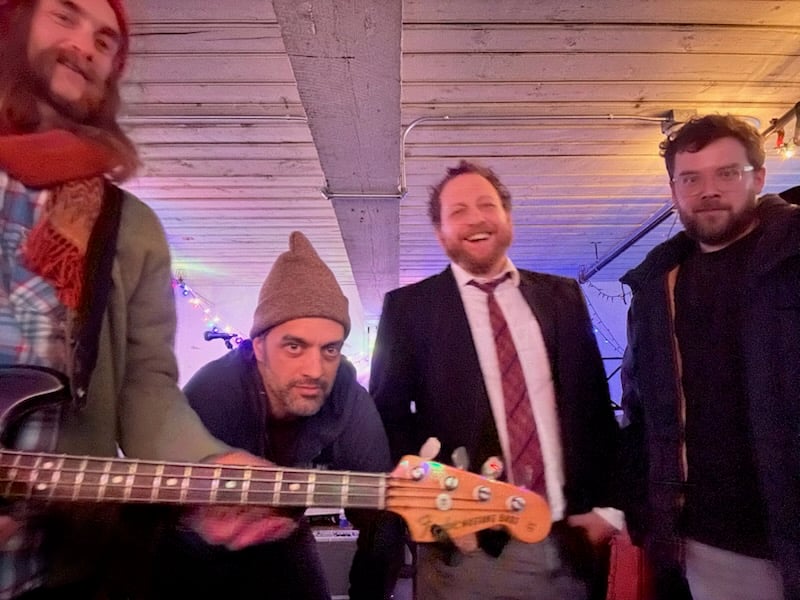 Four menstand side by side. One, on the left, is wearing a scarf wrapped around his neck and holding a bass guitar.