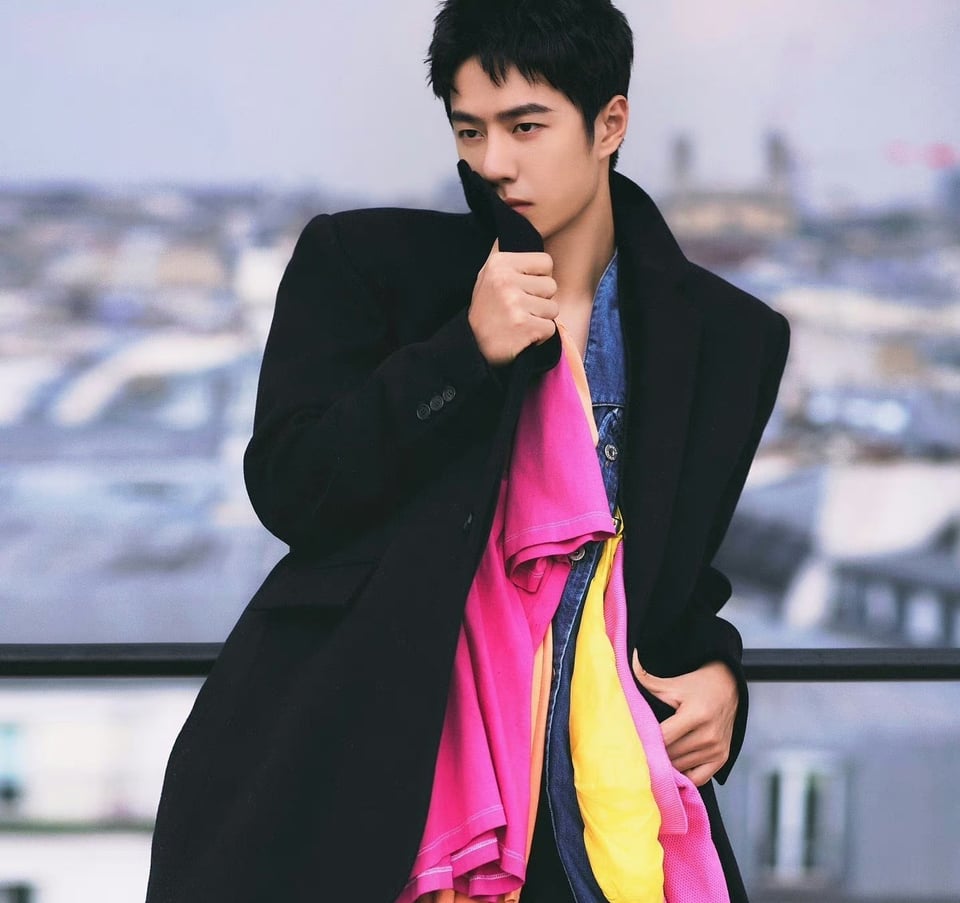 Wang Yibo wearing a fancy thick black overcoat over an inexplicable conglomeration of fleece towels in various bright colors, themselves draped like a weird scarf over a denim jacket. He's leaning against a railing in Paris looking effortlessly cool, despite being draped in literal rags.