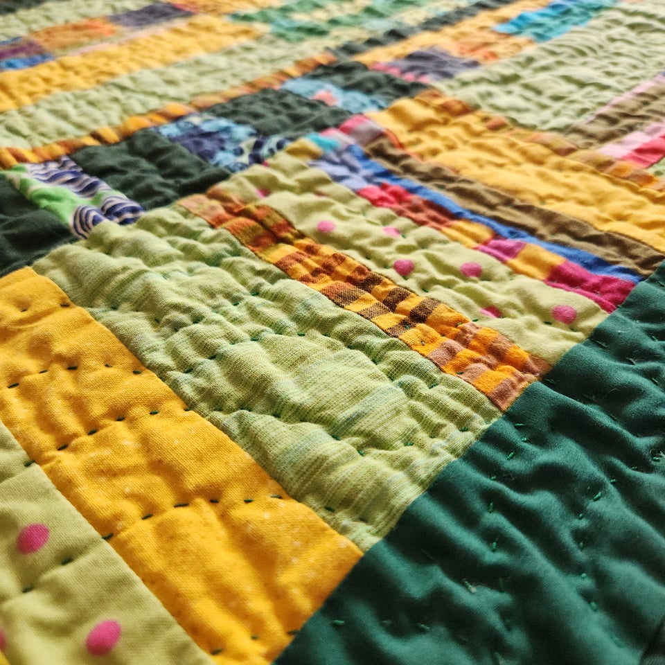 Close up photo of Sunshine Spreads a Carpet, showing fabrics in green, yellow, and other colors. The crinkly texture of the quilt and Sarah’s curved lines of hand stitching are visible.
