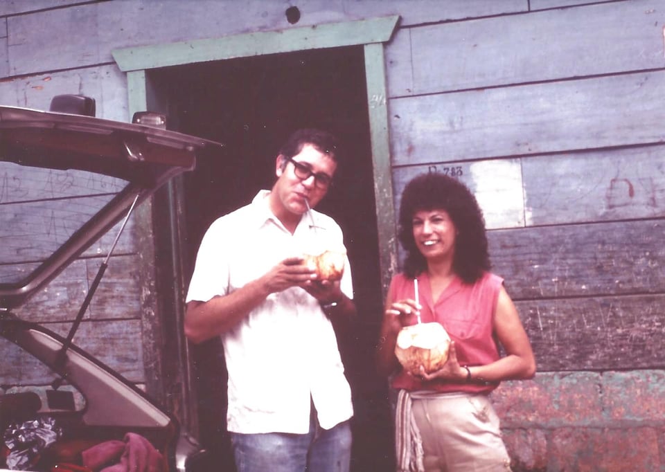 A young man and woman standing before a wood plank house drinking from coconuts.