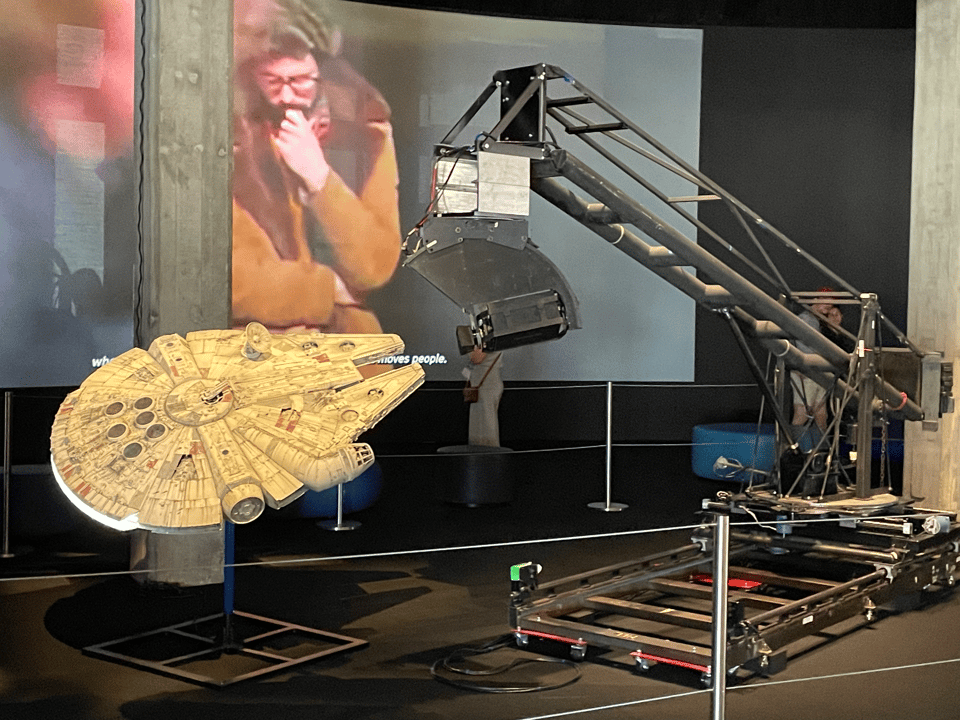 A large camera aimed at a spaceship model. On a screen in the background, young George Lucas stares at it all