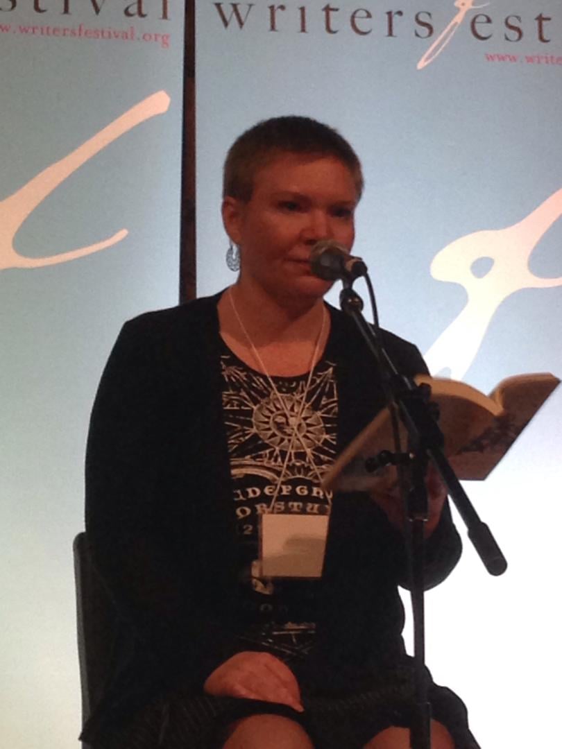 Kate in a ouija board top and buzz cut, seated and reading at a microphone.
