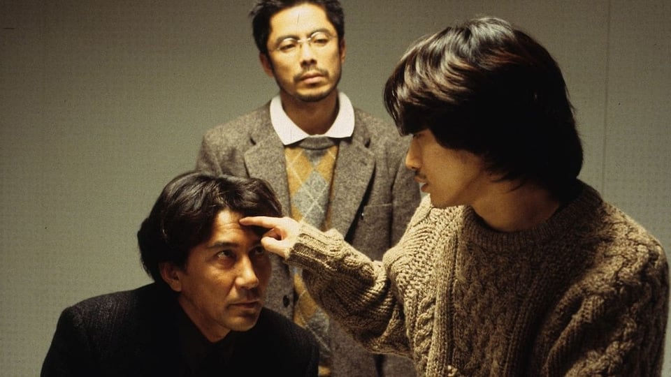 Three men in a plain-walled room: a seated man in a dark jacket looks up while a younger man in a cable-knit sweater points a finger at his forehead, and a bespectacled man in a tweed blazer and argyle sweater stands watching in the background.