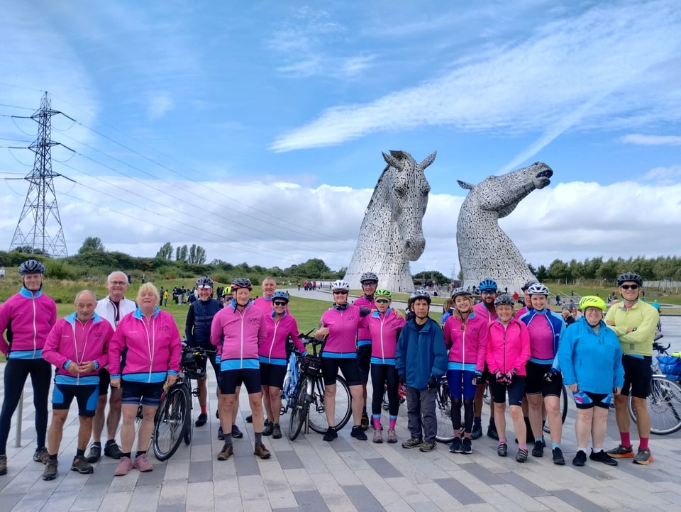 A group of around twenty people stand with bikes in front of the Kelpies statues, under blue skies. Many of them are in distinctive fluorescent pink jackets and most are wearing helmets.