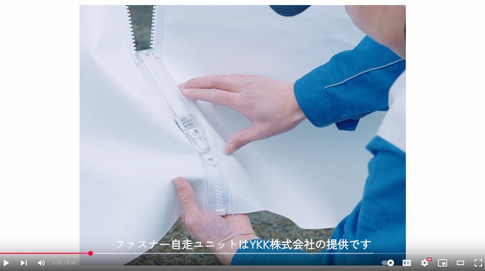 Screenshot of the YouTube video. It shows a close-up of the self zippering zipper, attached to two large plastic vinyl sheets. A man leans forward, his two hands touching it. The zipper is partly undone.