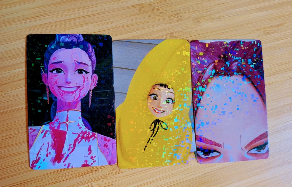 A photo of three Kpop Demon Hunters Photocards - photocards being the famous trading cards included in most Kpop albums of one or more (usually) idols either being cute or silly. This set are of the KPop Demon Hunters main characters - members of the group "Huntrix". Rumi, Zoey and Mira from left to right. Rumi is covered in demon effluence and smiling with a strained look on her face, based on a similar photocard from Kpop Idol Jeon Somi. The Second is Zoey, smiling and looking cute in a banana costume filter. Based on a real photocard of idol Park Chanyeol . The final is Mira, just her forehead, her eyes looking at the camera. Based on a photocard of idol D.O. from the group Exo. He had just taken a photo of his forehead, it's very funny.