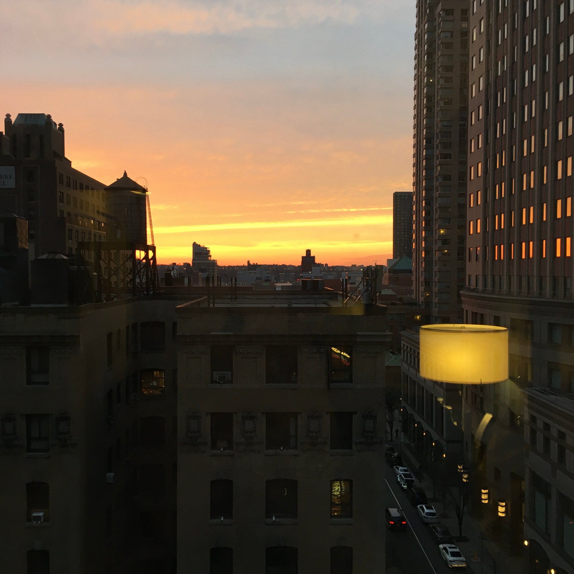 Photo depicts a sunset from a high-story building window. Other nearby buildings frame the bottom and right sides of the image. A reflection of an indoor lamp rests in the lower right corner.