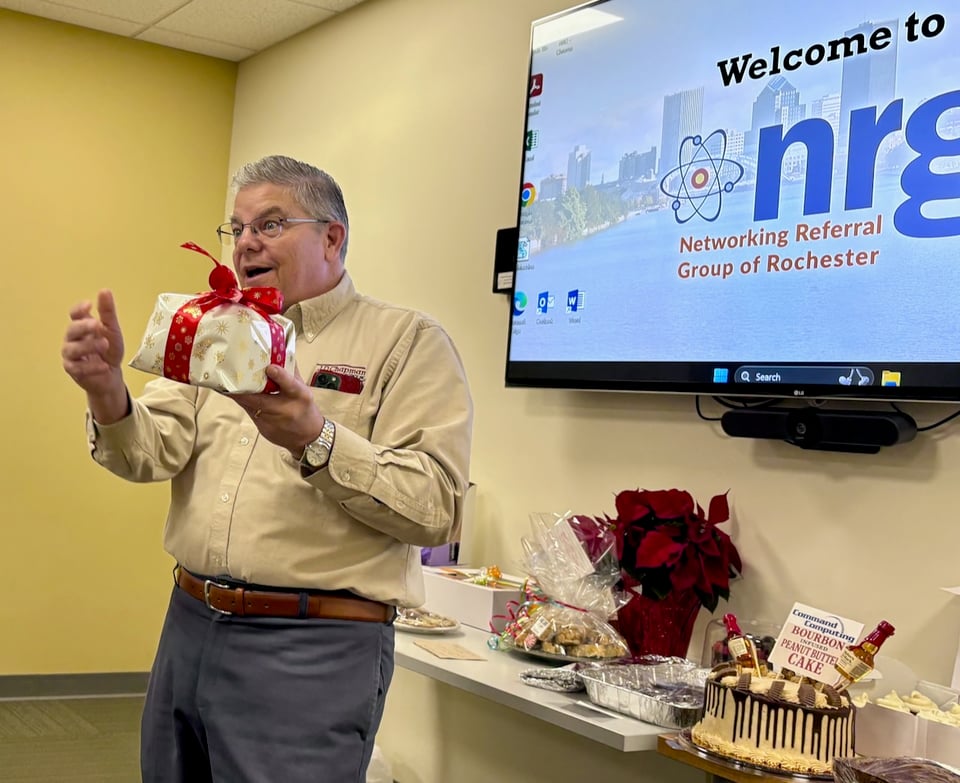 Bruce stands in front of the NRG welcome screen and table of baked goods while holding a gift-wrapped item. He’s gesturing and has a look of happy surprise on his face.
