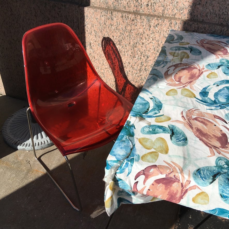Photo of a translucent red chair and a square table from an oblique, overhead angle. The table has a cloth with crab and seafood illustrations on it.