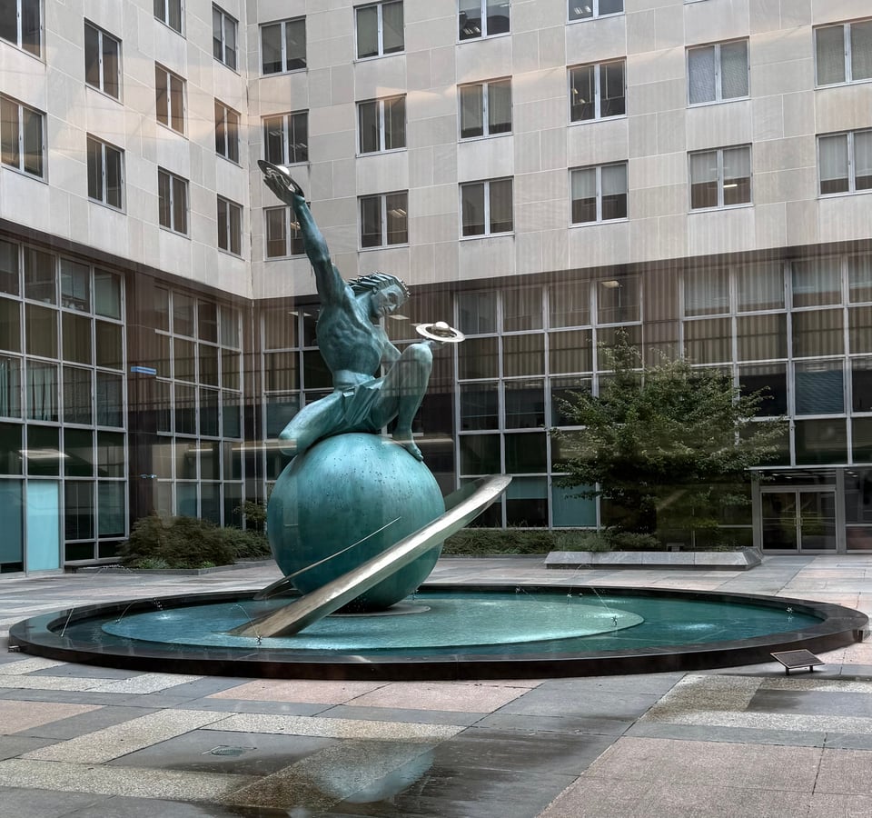 Photo of the Expanding Universe Fountain sculpture at the Harry S Truman State Dept. building, showing a bronze of a man crouching on a globe while he holds a pair of orbs with discs