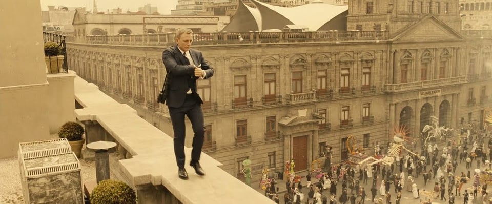 A still from the opening of Spectre (2015) with Daniel Craig's James Bond walking along a rooftop in Mexico City as a parade takes place below.