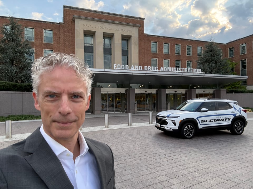 Selfie of middle-aged white man in dark gray suit and white dress shirt, standing in front of entryway for Building 1 of FDA headquarters. A security SUV is in the background