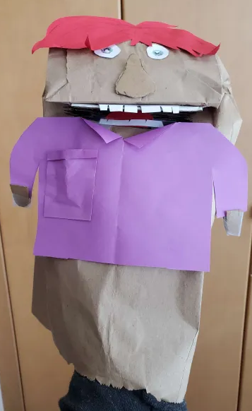 A paper bag puppet, with a shirt cut from coloured paper, red hair, and teeth.