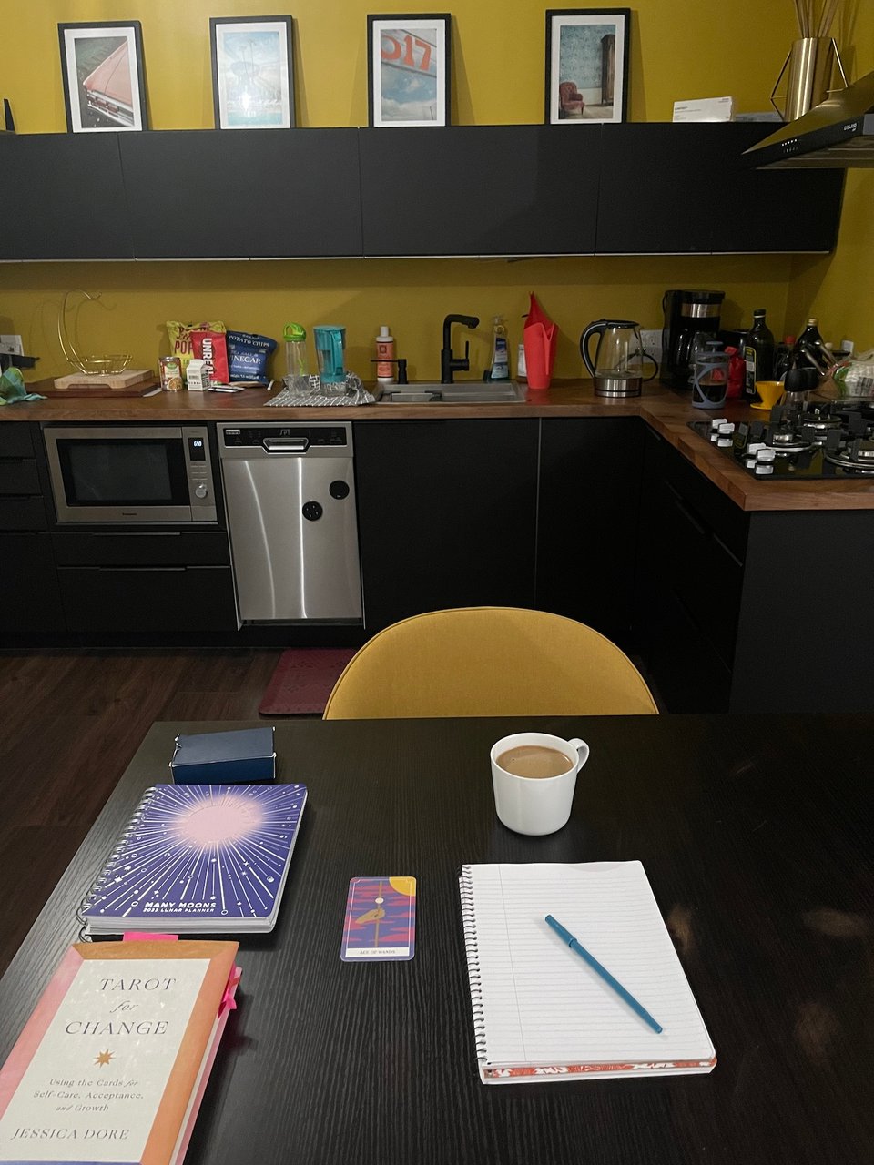 View of a table, with a notebook, a tarot card, a cup of coffee with cream, a moon notebook, and the book Tarot for Change by Jessica Dore. In the background is a yellow chair, and a kitchenette with mustard yellow walls.