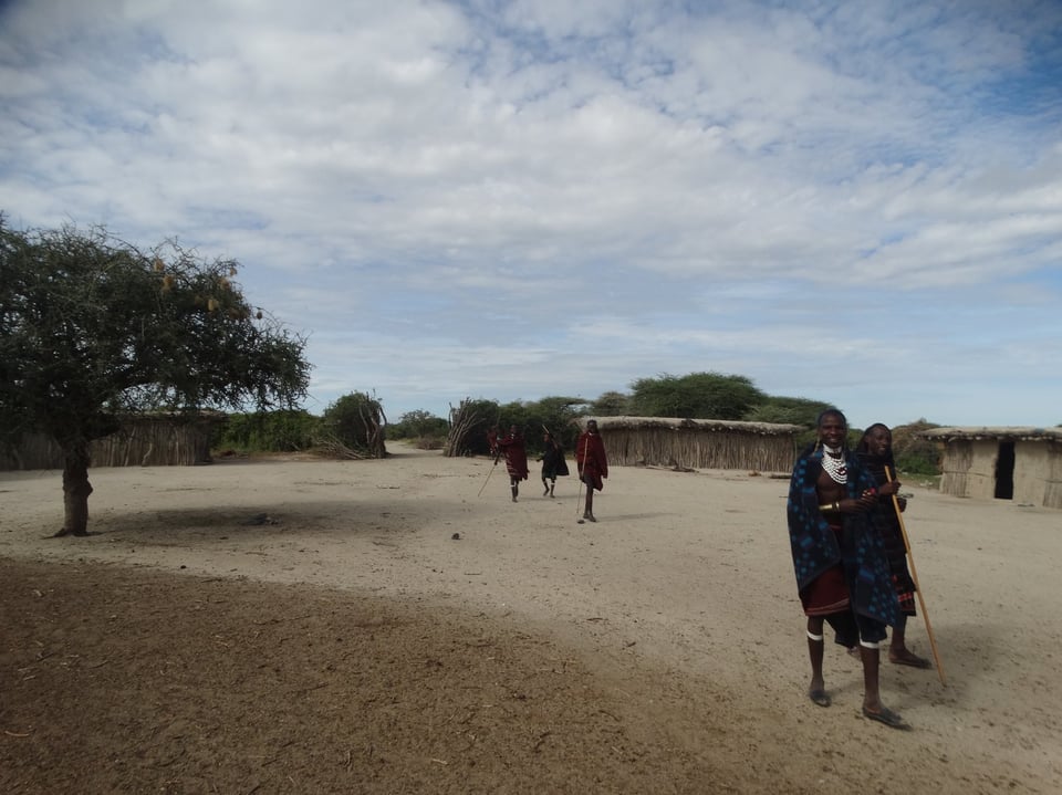Two Maasai men in traditional attire, carrying wooden sticks, pose for the camera while three others walk through their settlement in the background.