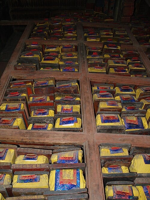 A tall wooden bookcase bearing manuscripts wrapped in yellow, blue, and red cloth stretches up into darkness.